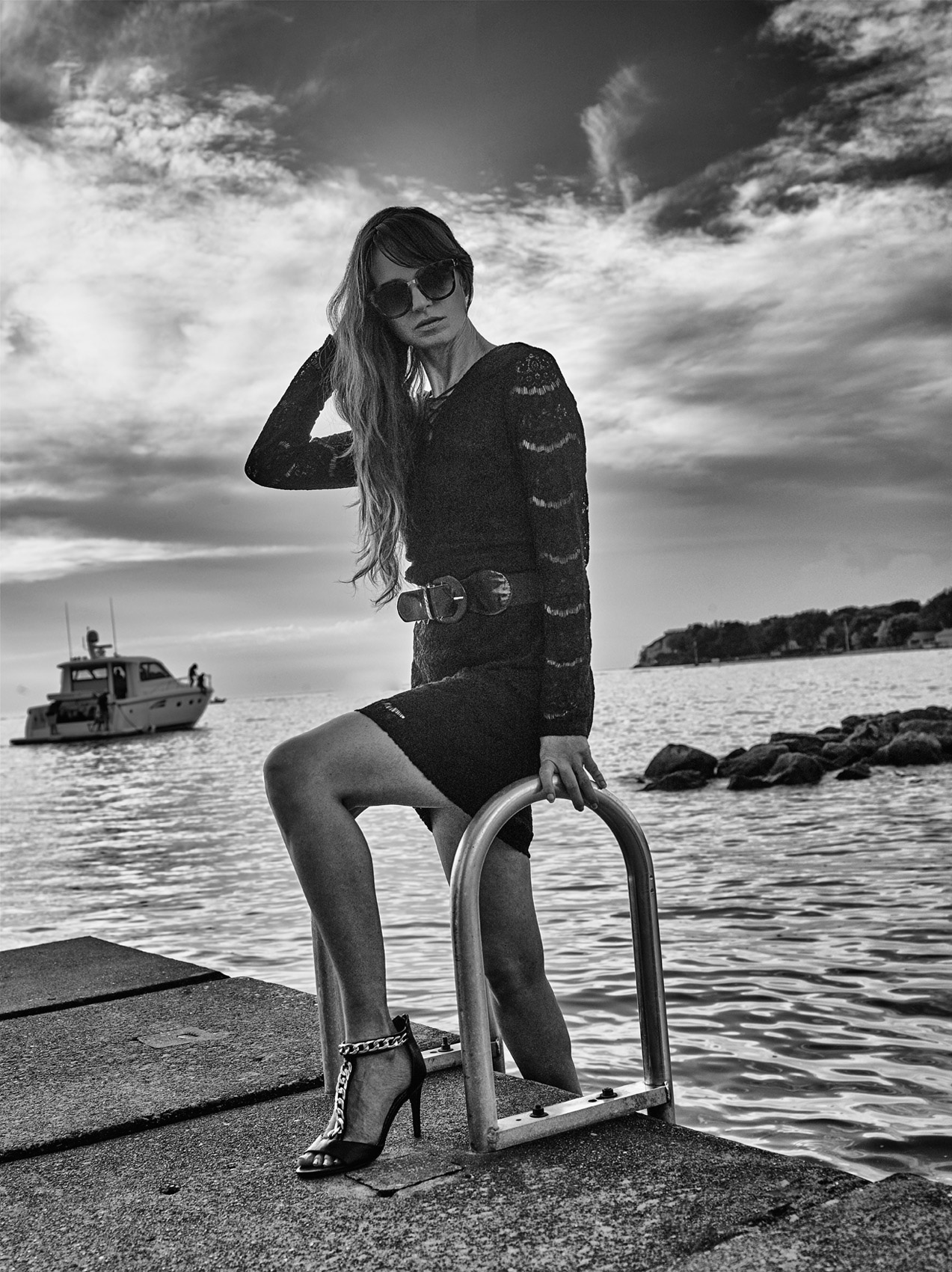  CASTAWAY_Model April In Black Dress & Sunglasses On Dock Ladder Holding Rail As Yacht Leaves Bay Island Rocks With Clouds Above Skyline At Catawba Island Country Club Pier 
