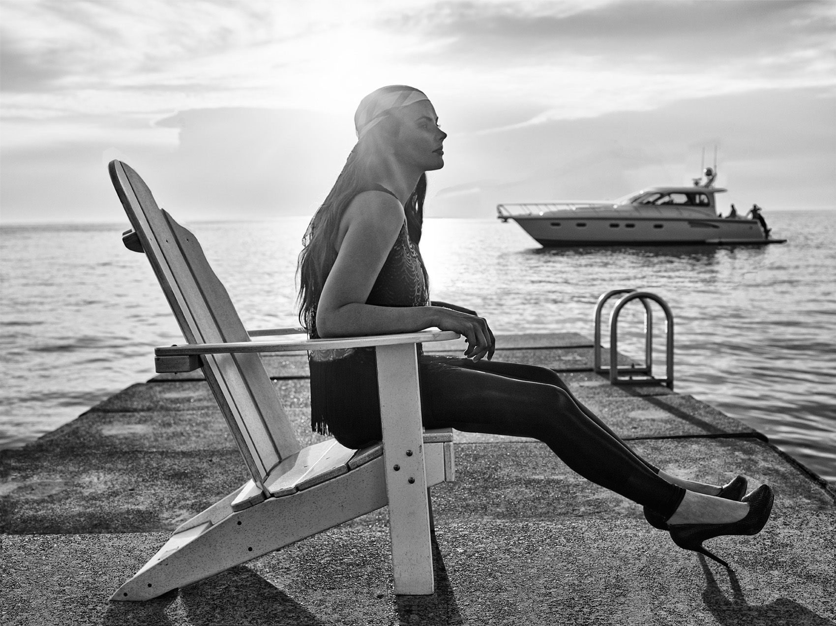 CASTAWAY_Model Elizabeth On Wooden Beach Chair With Legs Outstretched Across Pier Under Cloud Blanketed Horizon And Yacht Silhouetted On Water At Catawba Island Country Club 