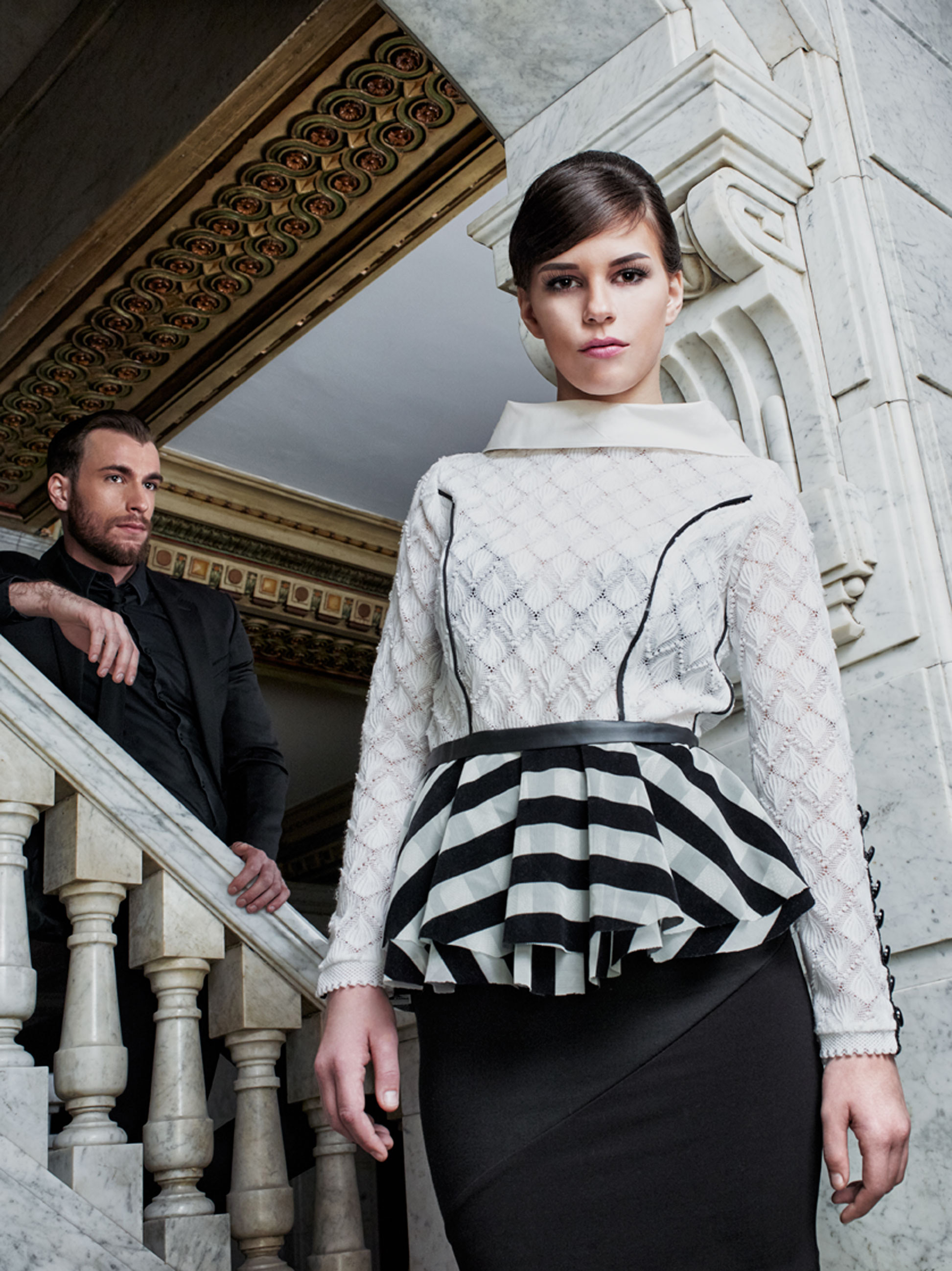 RULERS OF EVIL_7_Model Morgann Portrays An Evil System Dressed In White & Black Top With Black Skirt Staring Down At Viewer In Front Of Marble Stairway With Model Jon Portraying An Evil Henchman Perched Above On Stairs Surveying Hall In Background In Columbus Ohio 
