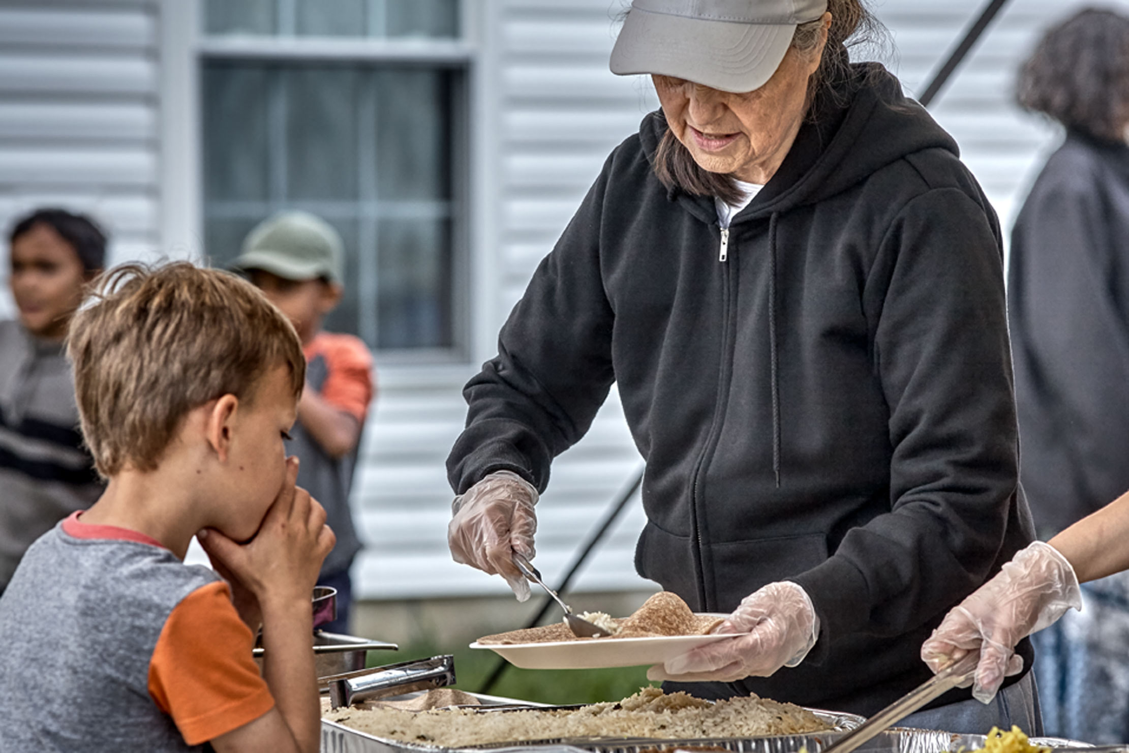  Lifestyle Portrait At West Salem Mission Campmeeting  Little Boy Wearing Orange And Gray Tee Shirt Awaits Older Woman In Gray Cap And Black Sweat Shirt Preparing Food Plate
