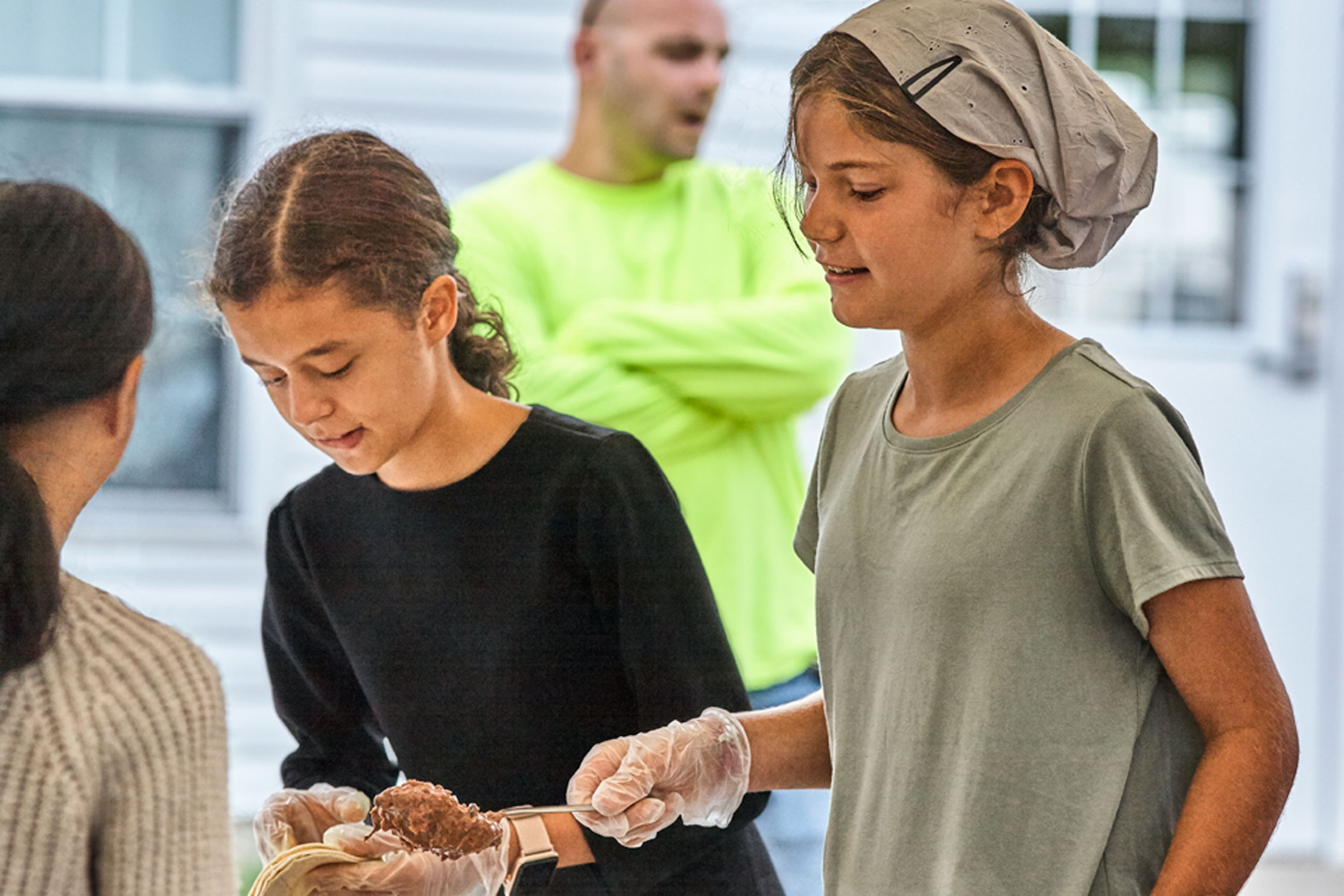   Lifestyle Portrait At West Salem Mission Campmeeting With Girl Wearing Light Green Shirt And Head Scarf Talks To Girl In Front Of Her While Girl Flanking Prepares Plate Using Bokeh To Enhance Scene 